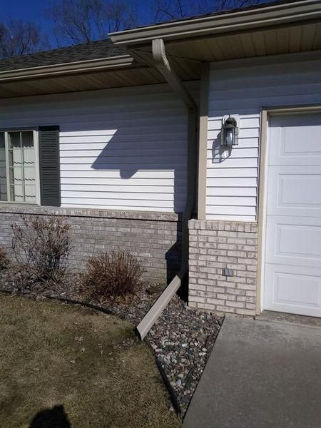 A residential house exterior showing a white garage door, siding, brick foundation, and a downspout draining.