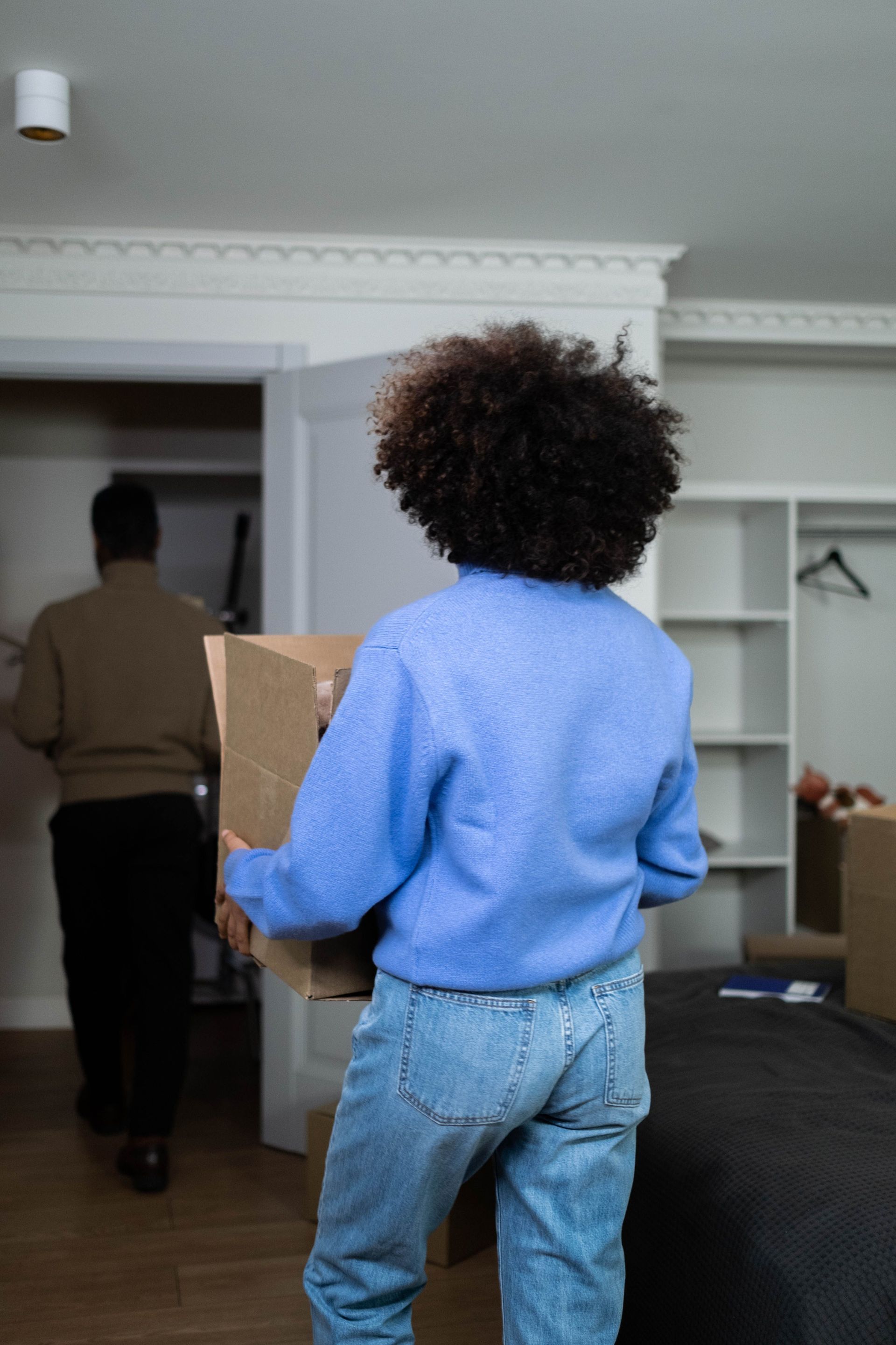 Person carrying a cardboard box in a bedroom with an open closet and another person at the doorway