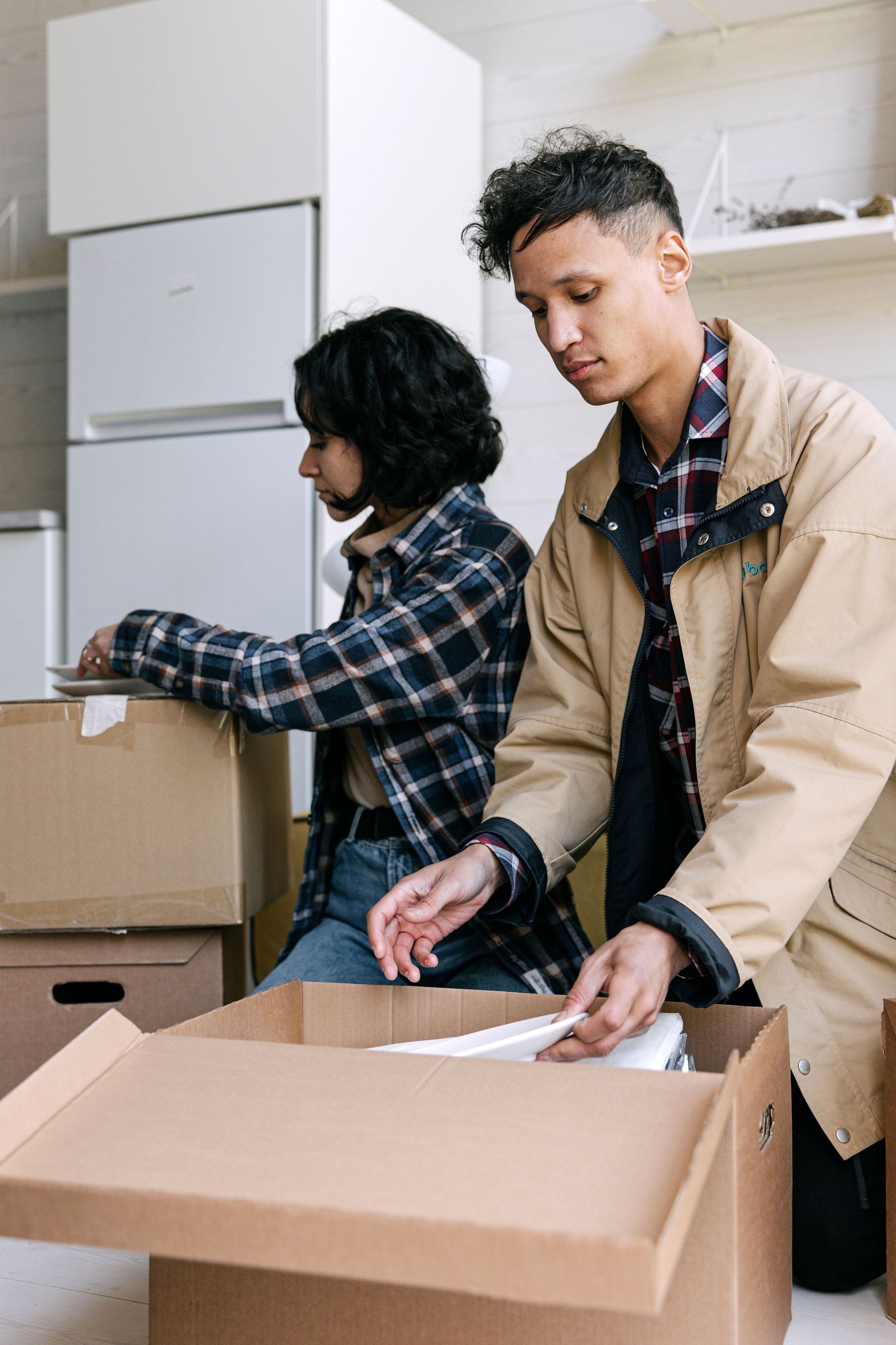 Two people unpacking boxes in a bright room, one folding a cardboard box and the other moving items.