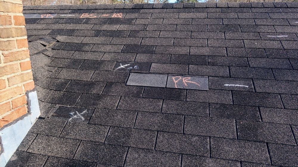 Dark shingled roof with several damaged or patched shingles near a brick wall.