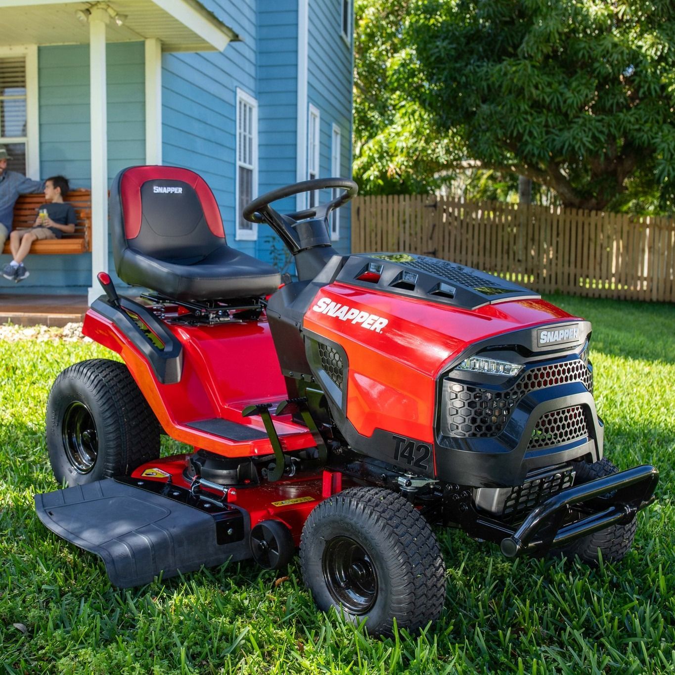 Red Snapper lawn tractor on grass beside a blue house, with a leaf sweeper attachment.