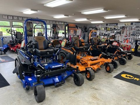 Row of blue, orange, and black riding lawn mowers displayed in a showroom, with tools on the walls behind them.