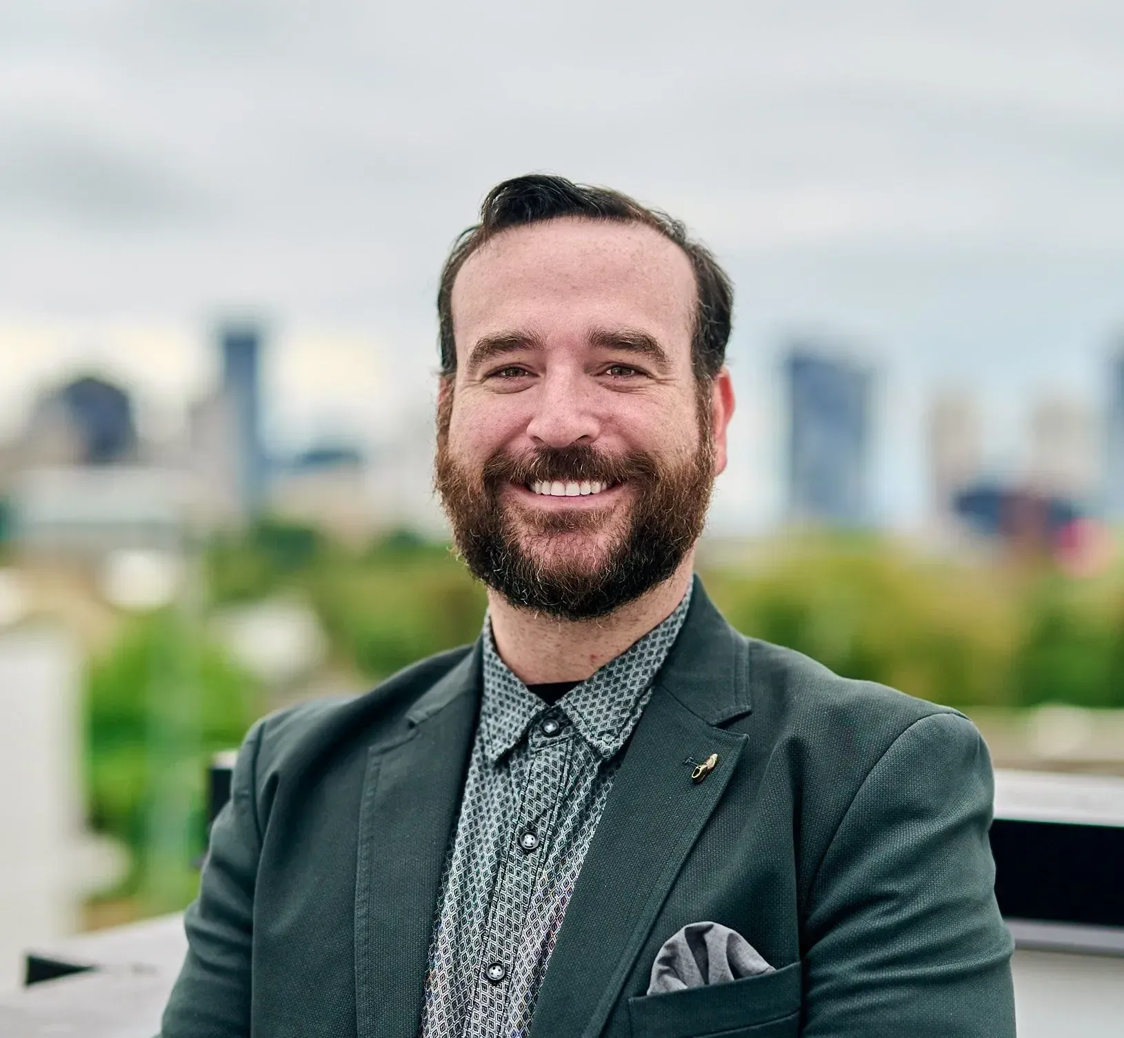 Man with beard smiles, wearing a patterned shirt and green blazer, against a city backdrop.