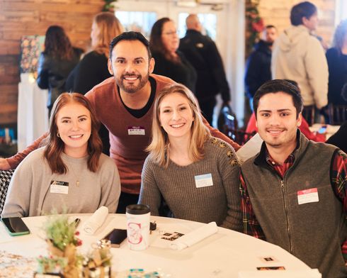 Four people smiling at a table at an event. Other people are in the background, festive decorations.
