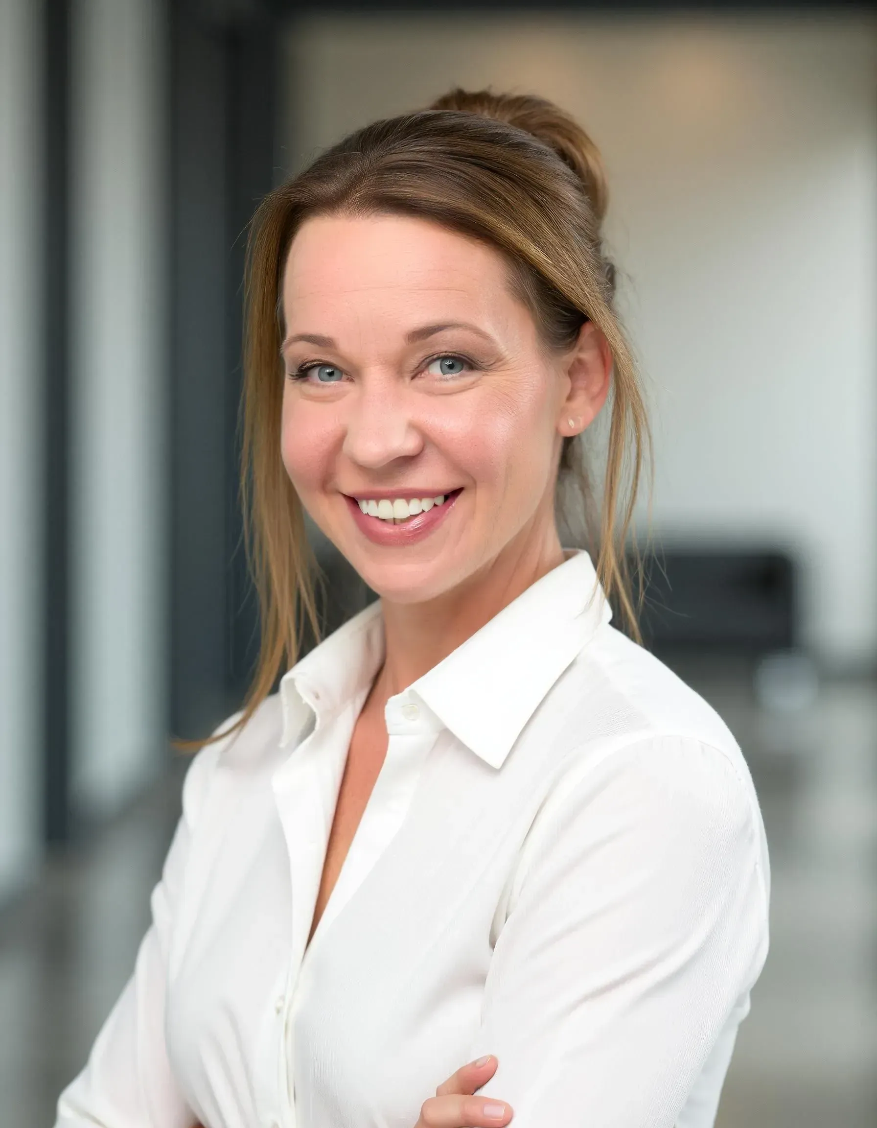 Woman in white shirt smiles at the camera, light brown hair pulled back, blurred background.