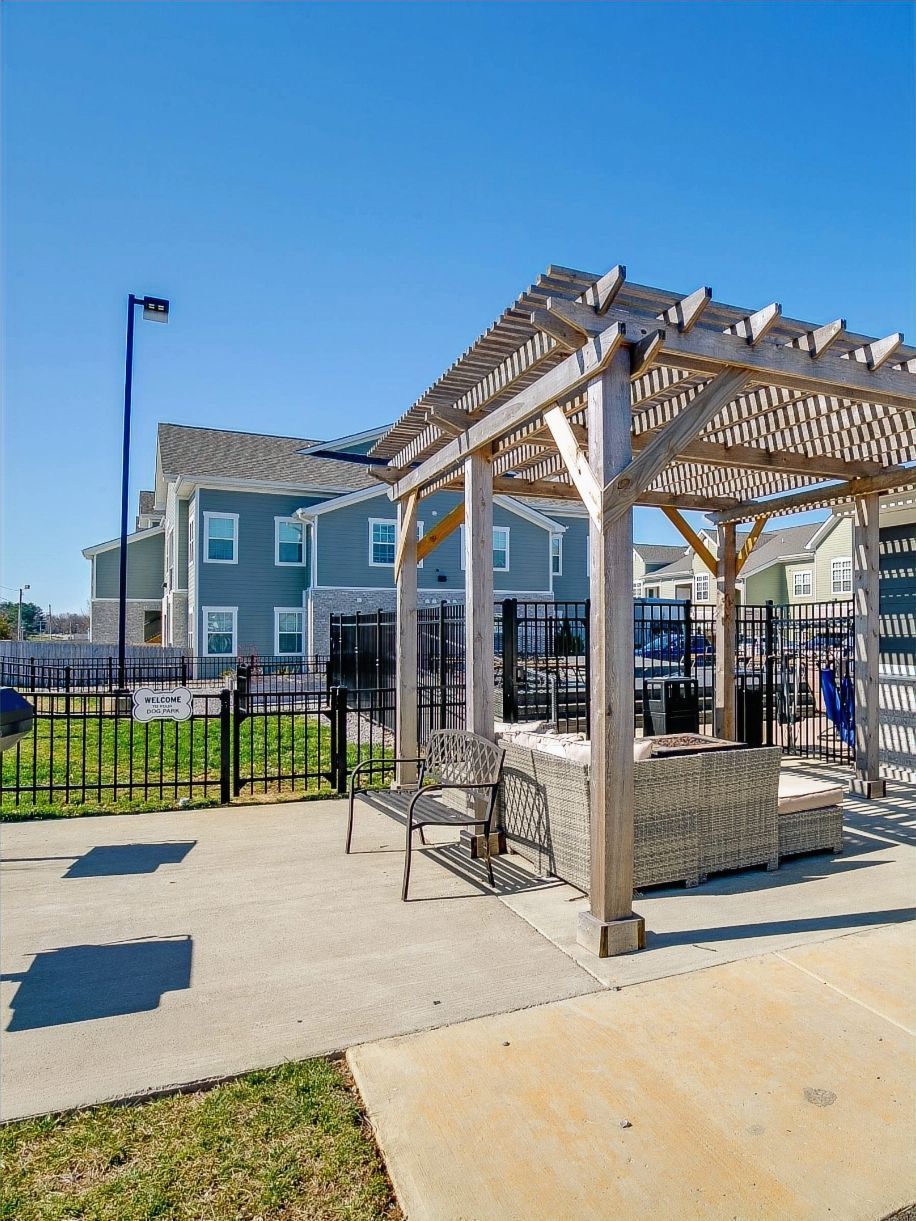 Wooden pergola with seating area, fence, and buildings under a blue sky.