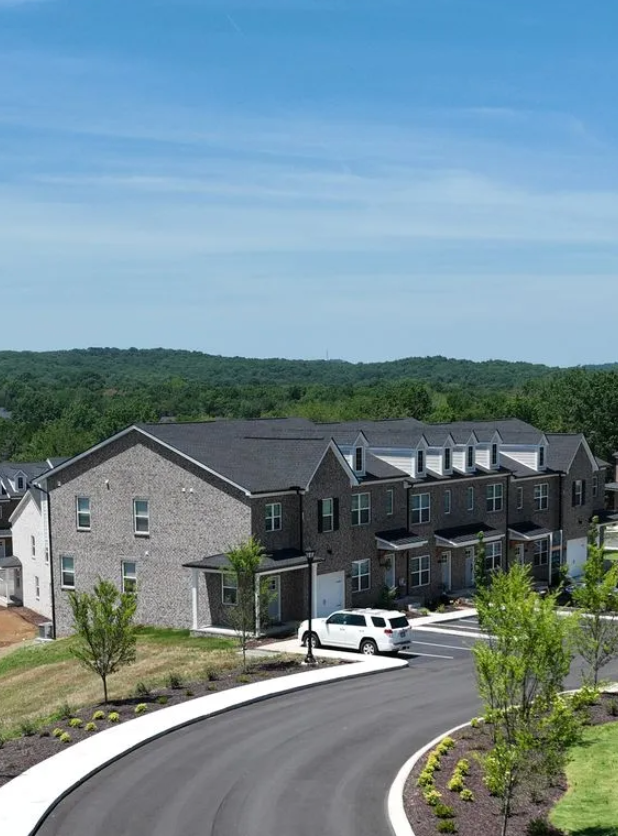 Townhouses with grey brick exteriors, asphalt road, blue sky, and green trees.