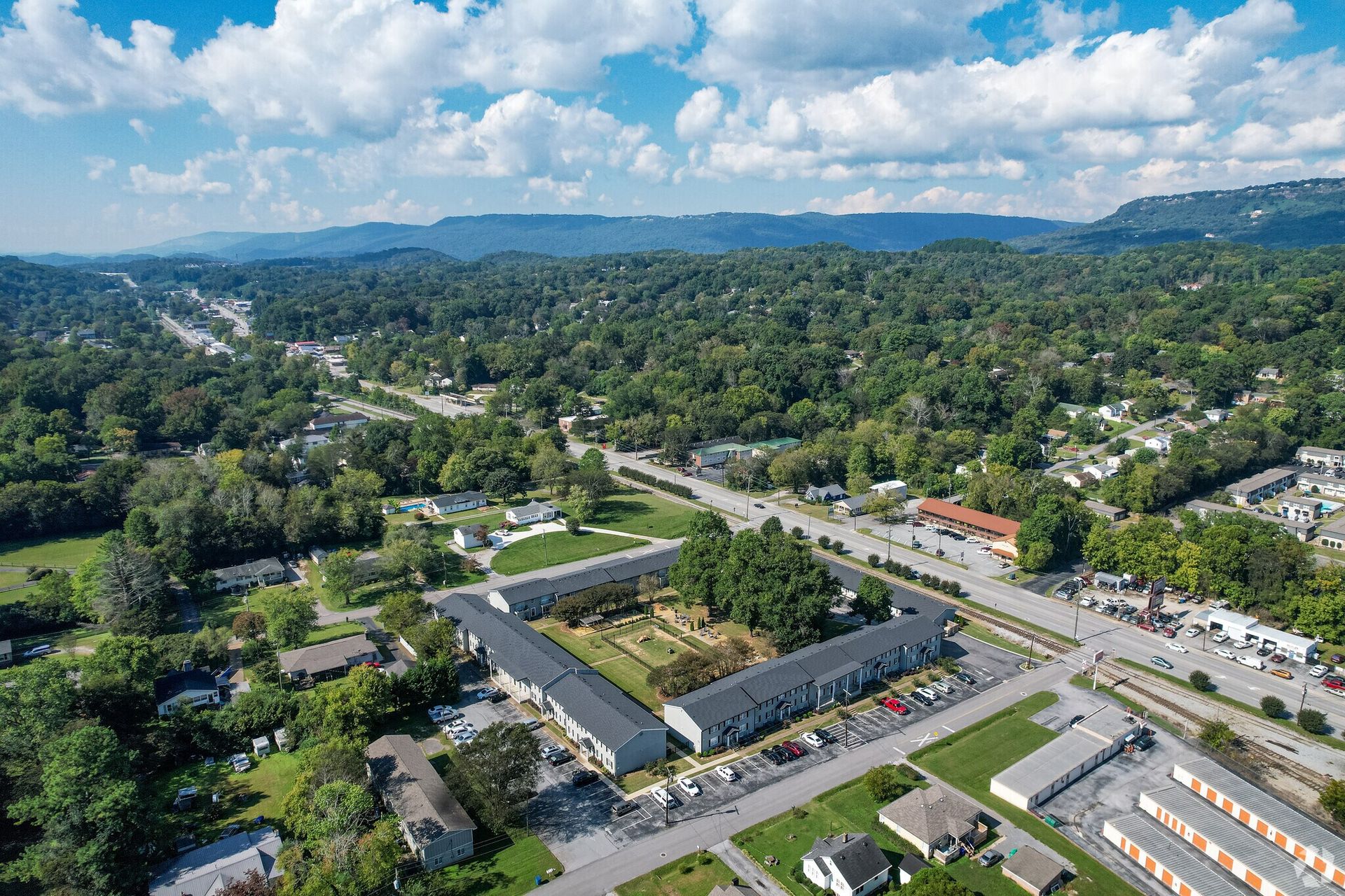 Aerial view of apartment complex surrounded by trees, roads, and mountains under a blue sky.