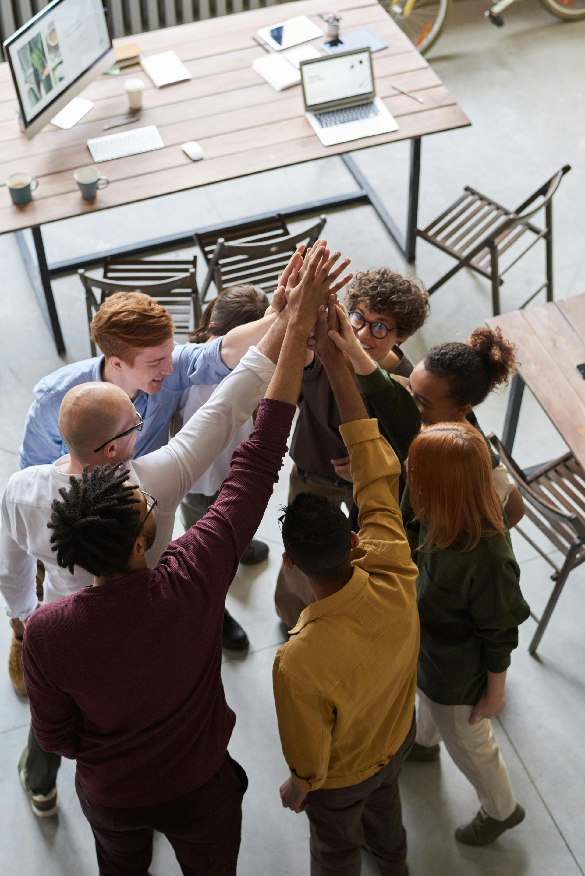 Group of people high-fiving in a modern office setting. Hands raised in center, wooden tables around.