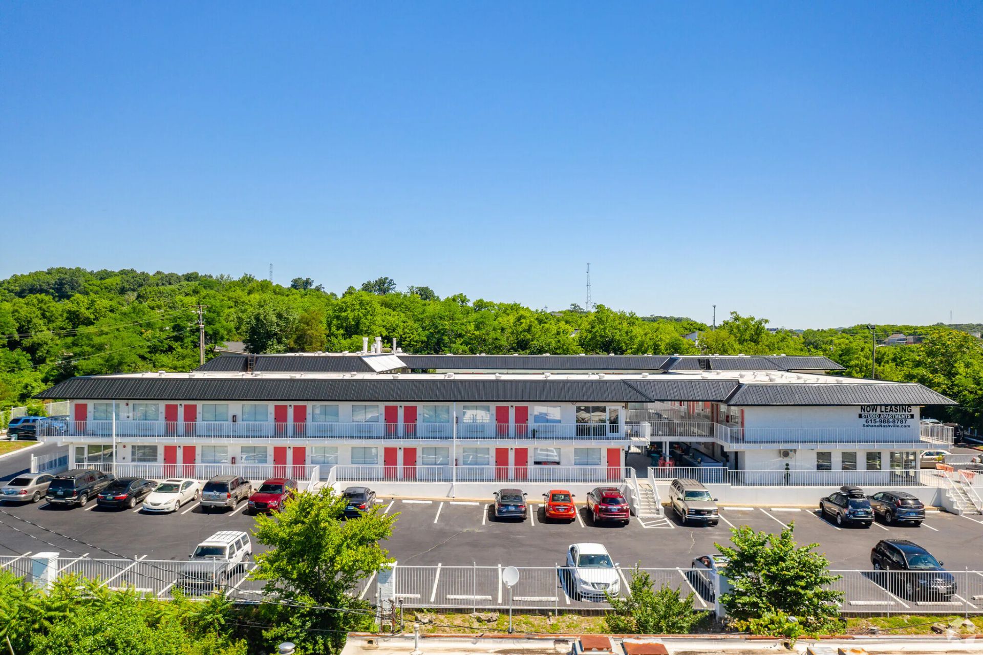 Motel exterior, two stories, red accents, white facade, parking lot with cars, green trees, and clear blue sky.