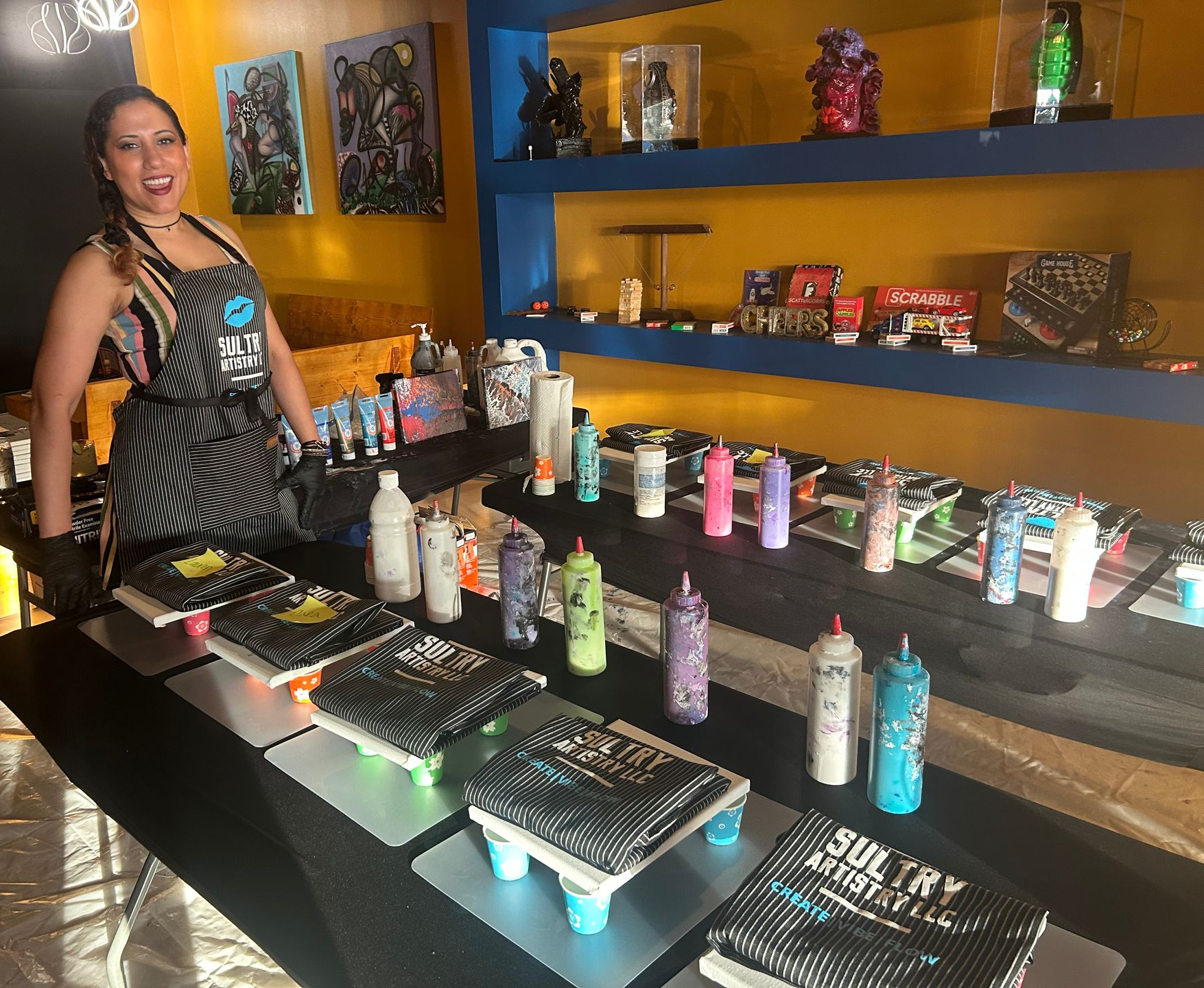 A smiling woman in an apron stands behind a table displaying aprons, canvases & rows of paint bottles.