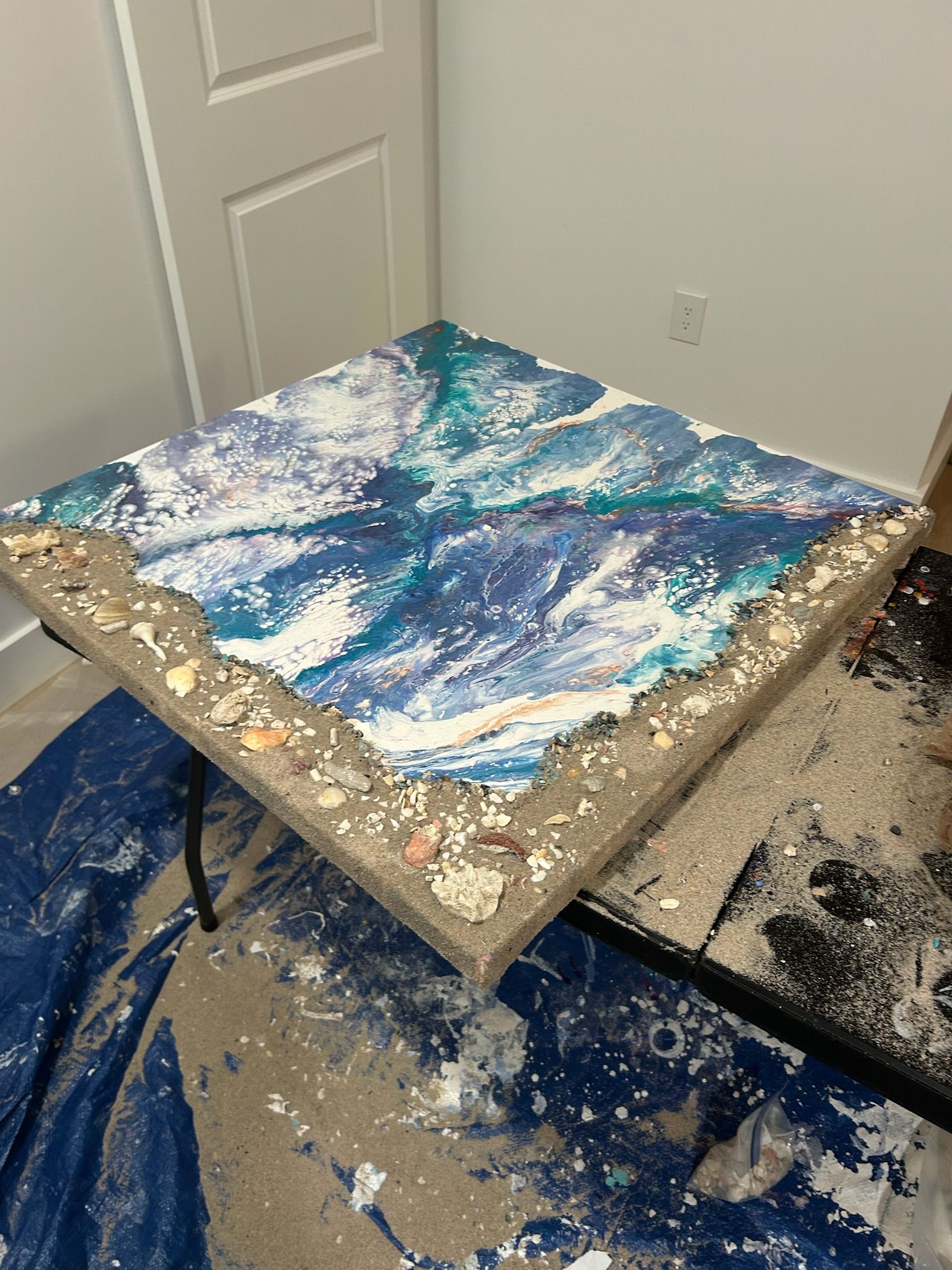 A square table features an abstract blue and white ocean-inspired acrylic pour painting surrounded by a textured sand border.