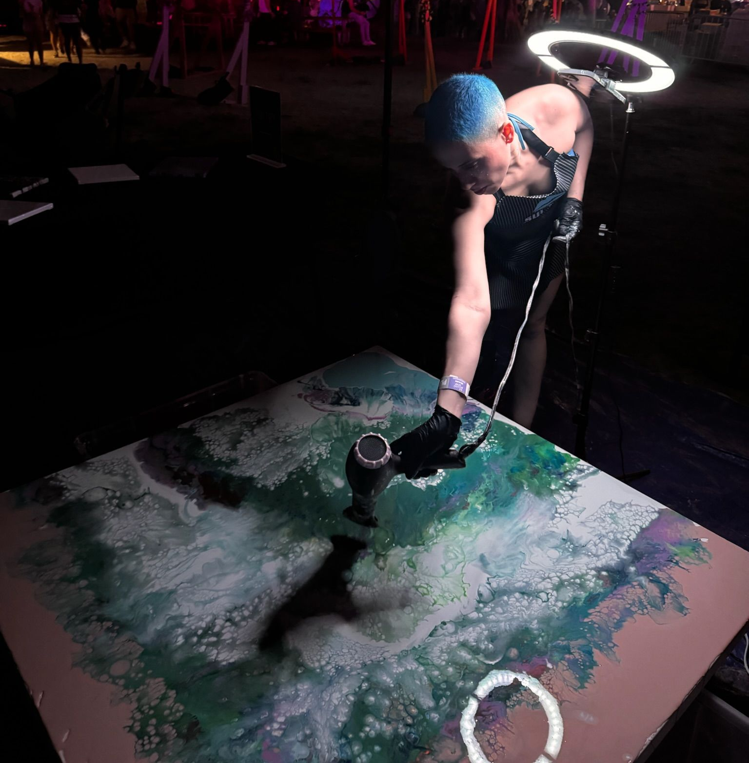 An artist with blue hair uses a heat gun to manipulate wet paint on a large canvas under a ring light in a dark space.