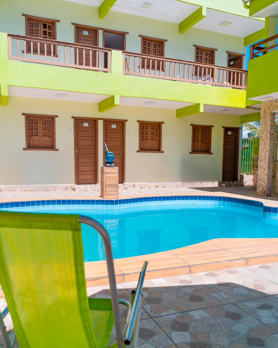 Piscina em um pátio, ladeada por um prédio de dois andares com detalhes em verde, e uma cadeira verde-limão em primeiro plano.