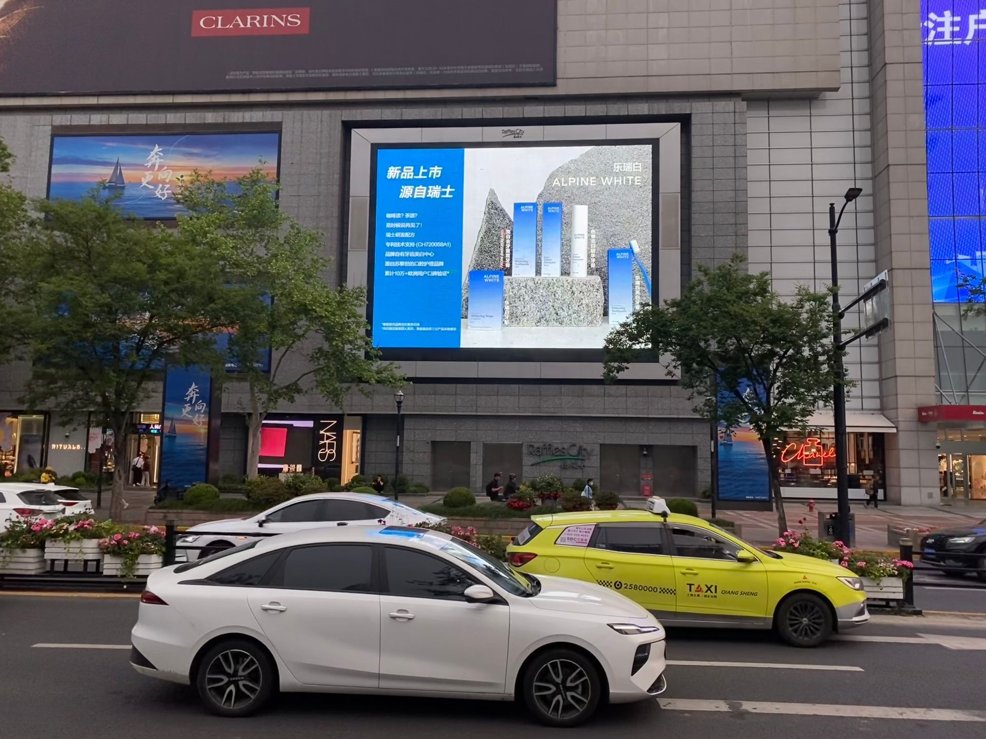 City street with a large digital billboard and several cars passing by, including a white sedan and yellow taxi.