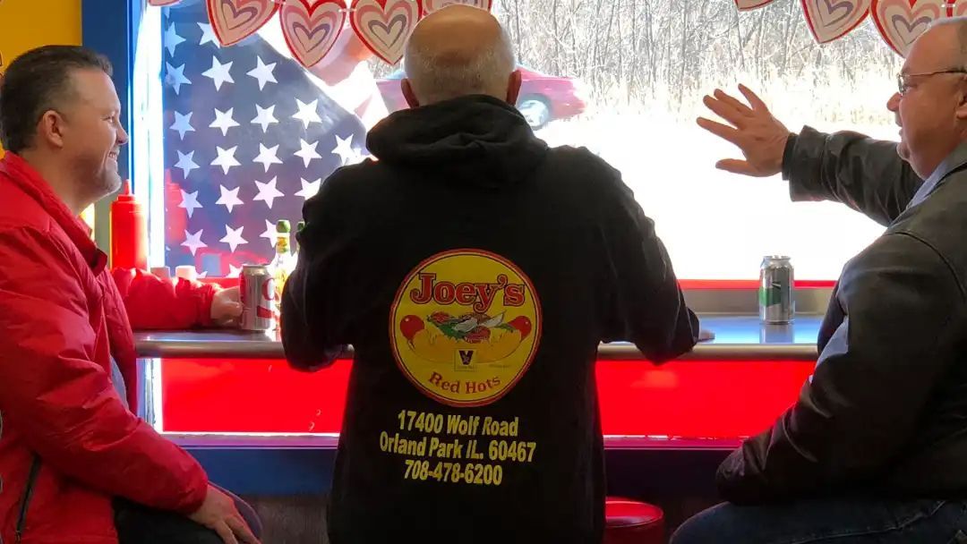 Three men at a diner counter with an American flag visible through the window. One man wears a hoodie with a restaurant logo.