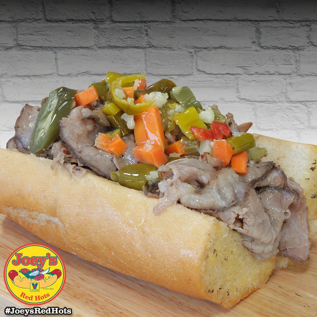 Sub sandwich on a wooden board, topped with sliced meat and pickled vegetables.
