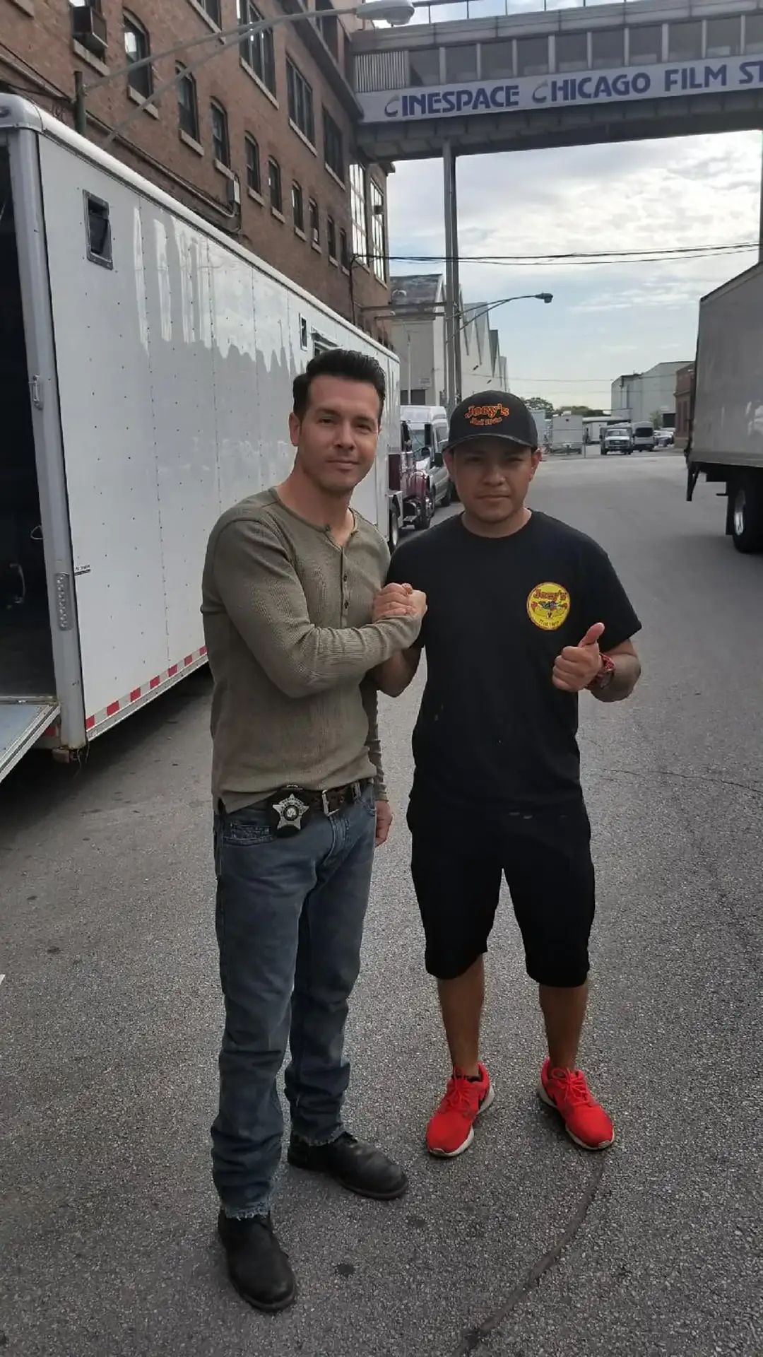 Man in jeans and man in black shirt shaking hands outdoors, near a white trailer.