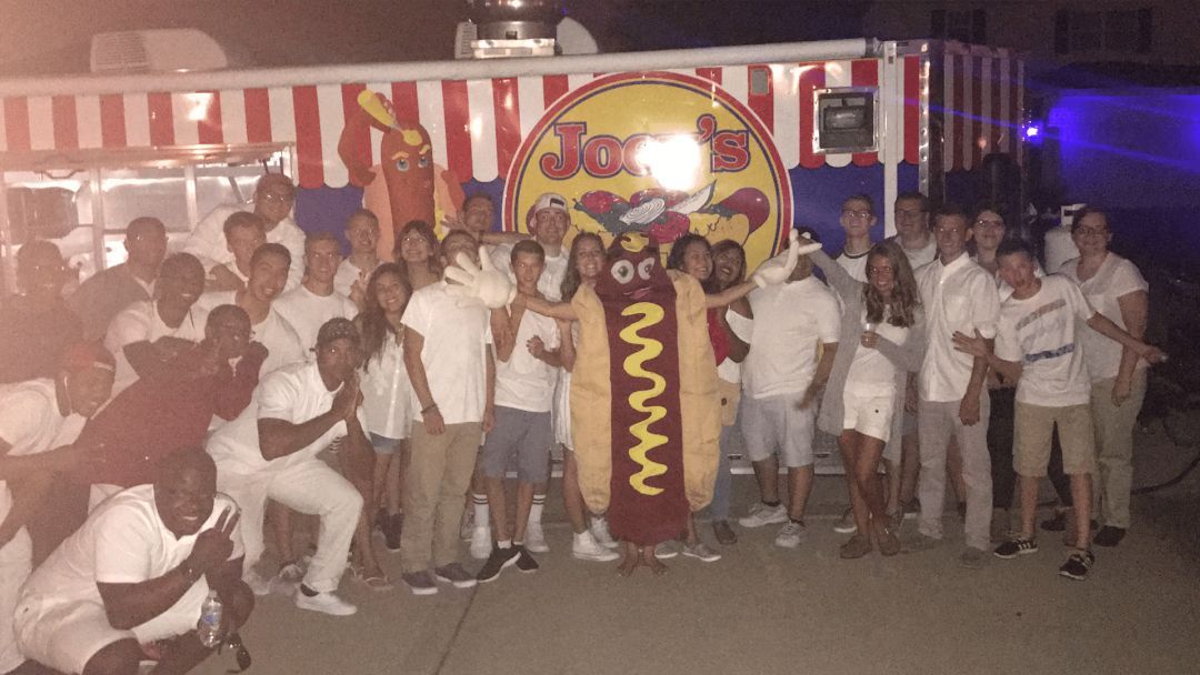 Group of people in white clothing pose with a hot dog mascot in front of a food truck with