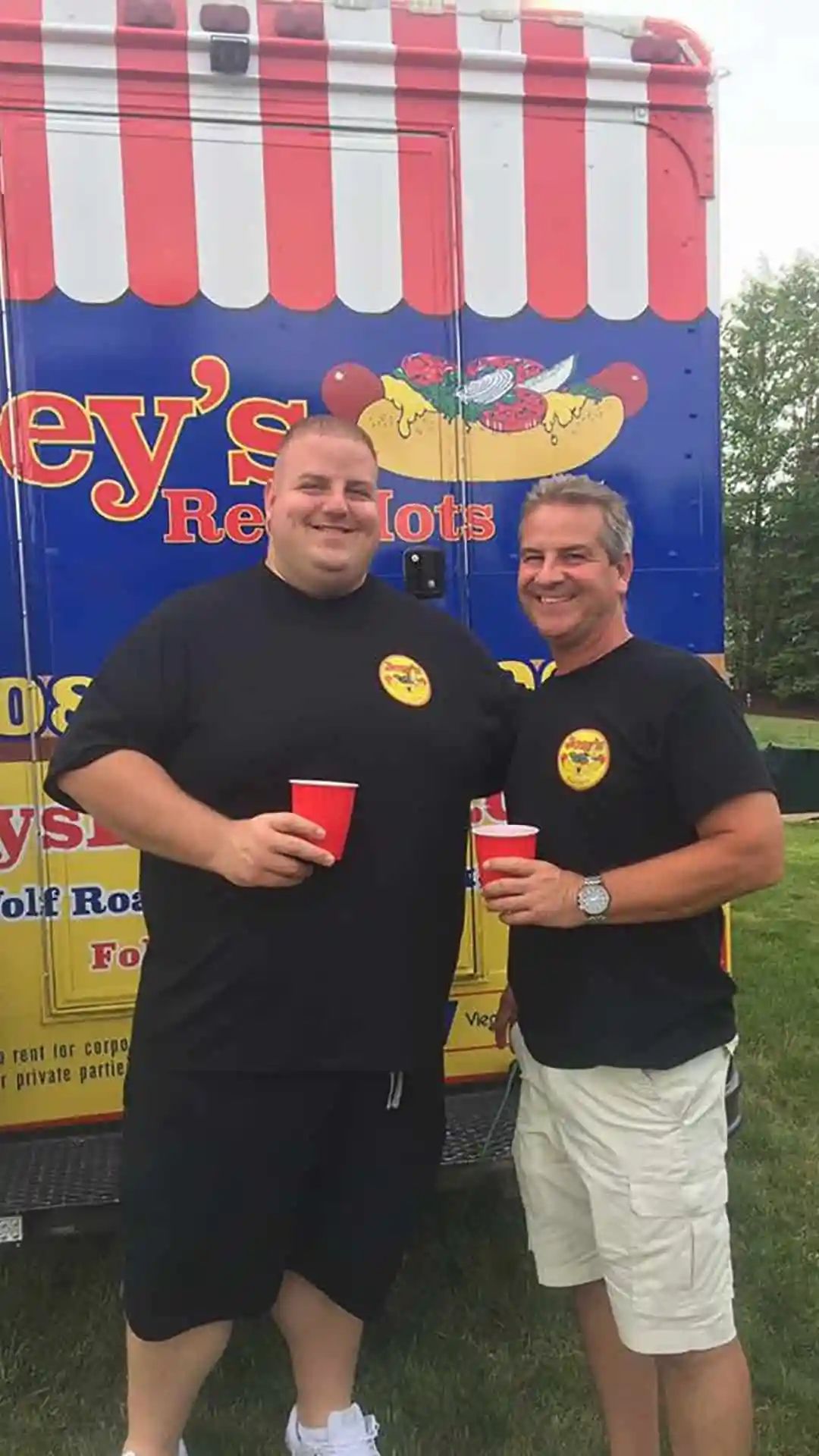Two men stand in front of a hot dog truck, each holding a red cup, smiling.