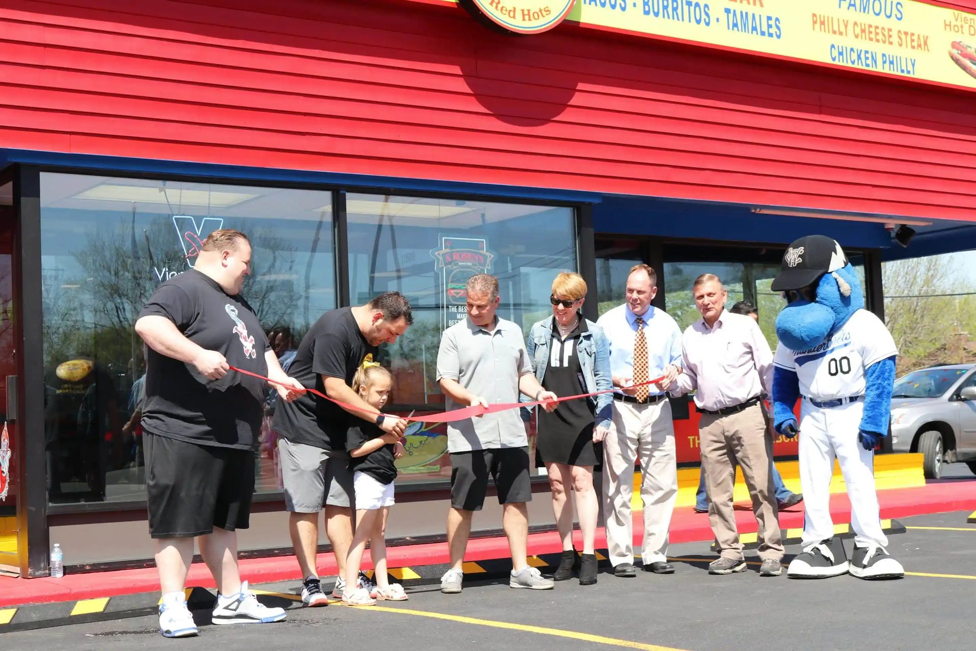 People cutting a ribbon in front of a red building, possibly a restaurant. A mascot stands at the end.