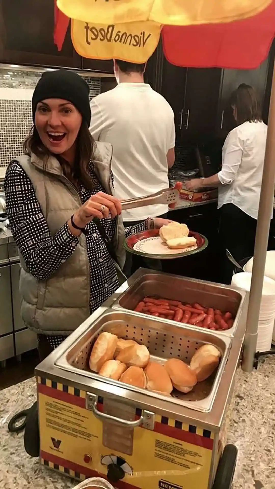 Woman with tongs at a hot dog cart, smiling. Hot dogs and buns in steamer, others behind.