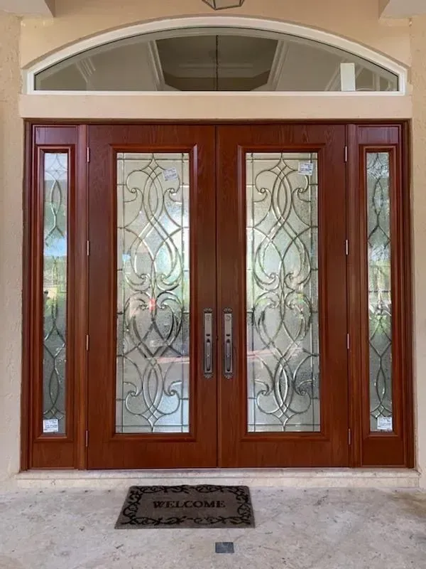 Wooden double front doors with frosted glass panels and a welcome mat beneath a curved transom window
