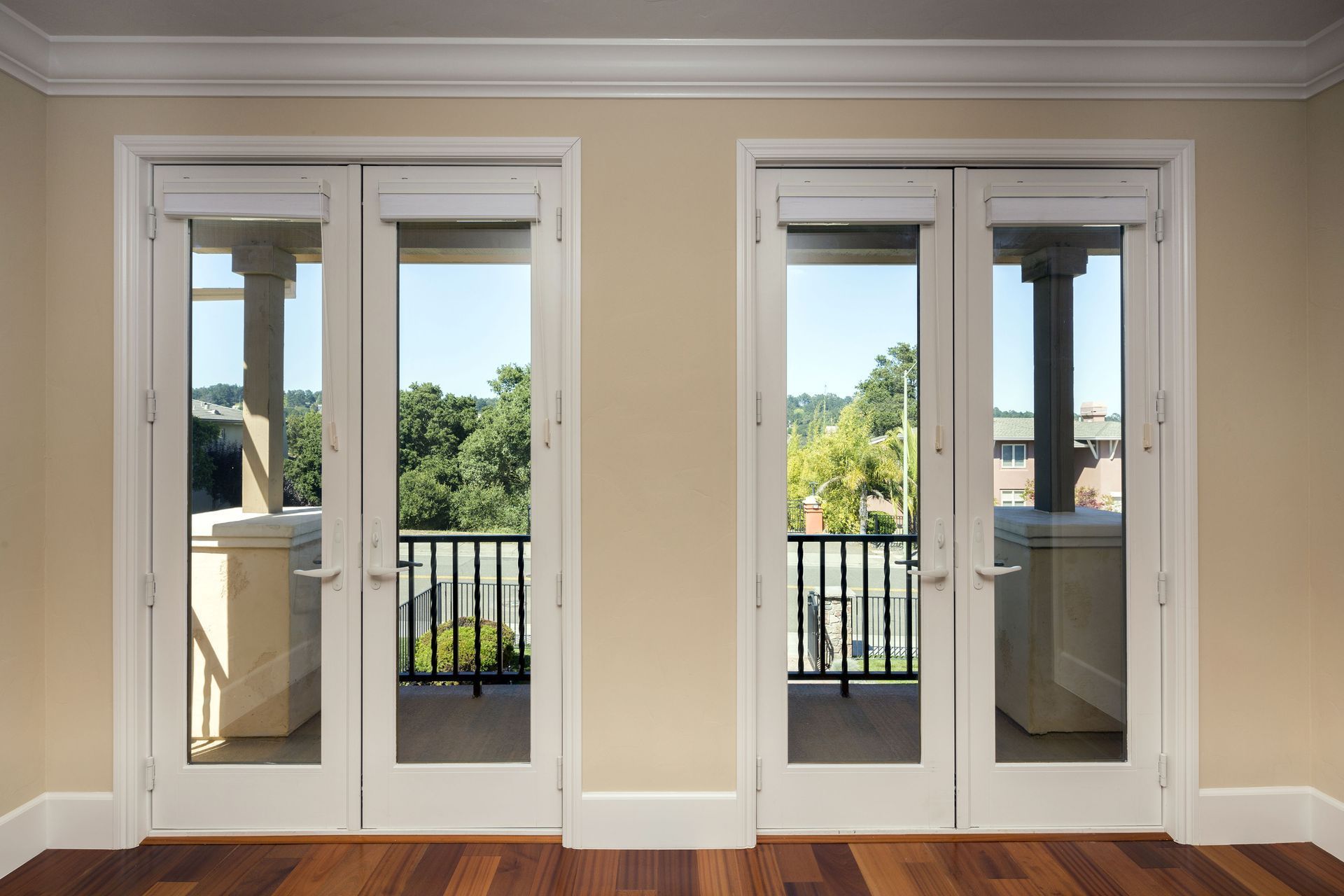 Two white French doors in a beige room with wood floors, opening to a balcony and green trees outside