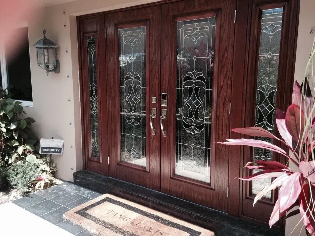 Front entrance with dark wood double doors, glass panels, and a doormat beside potted plants