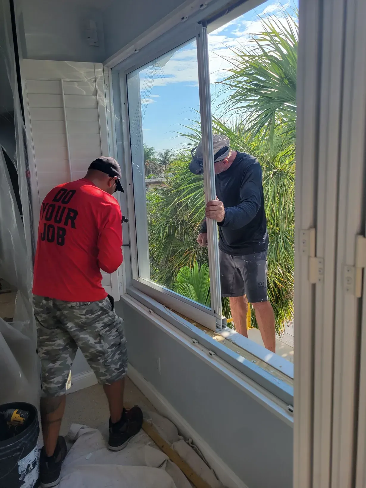 Two men working to install a large window inside a bright room while one stands outside holding the frame.