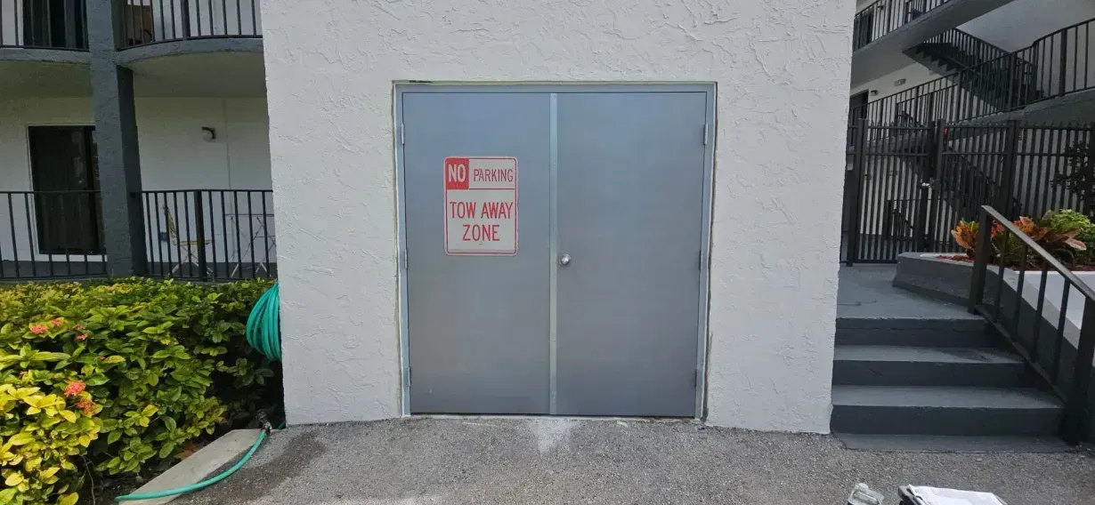 Gray double metal door in a concrete wall beside a stairway and shrubs, with a warning sign posted on it