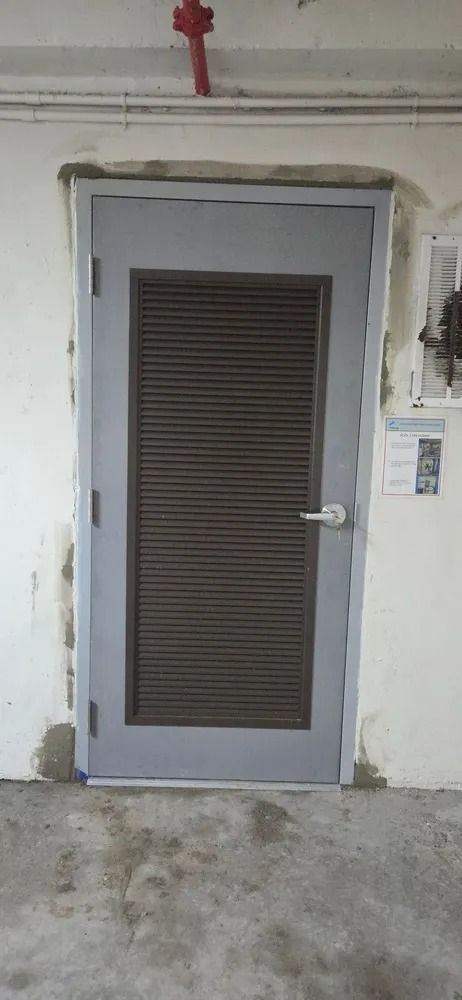 Gray door with a dark louvered window in a white wall, with a small keypad panel beside it.