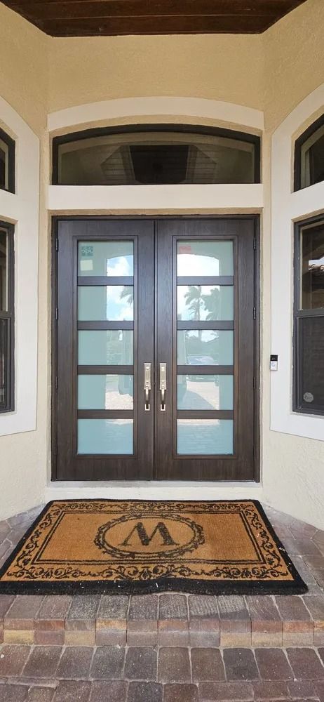 Front entrance with dark double glass doors, transom windows, and a patterned doormat on a brick porch.