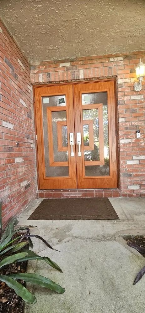 Brick entryway with wooden double doors, glass panels, and a doormat under a covered porch.