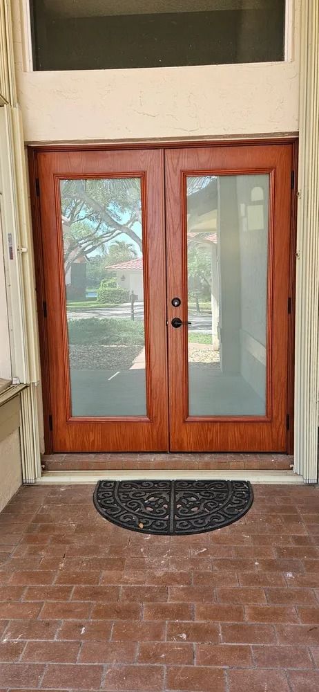 Wooden double front doors with glass panels and a black semicircle doormat on a brick porch