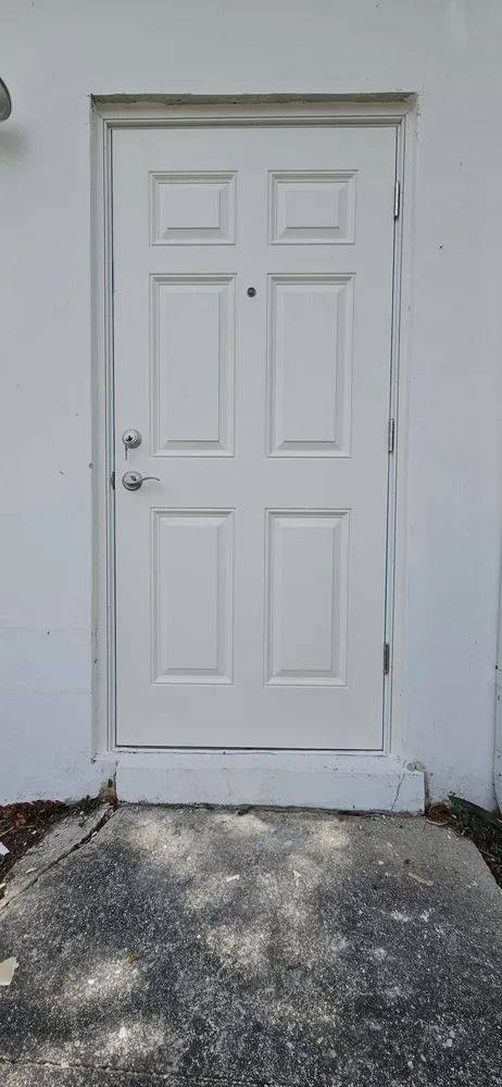 White exterior door with four panels and silver knob in a plain wall above a concrete step.