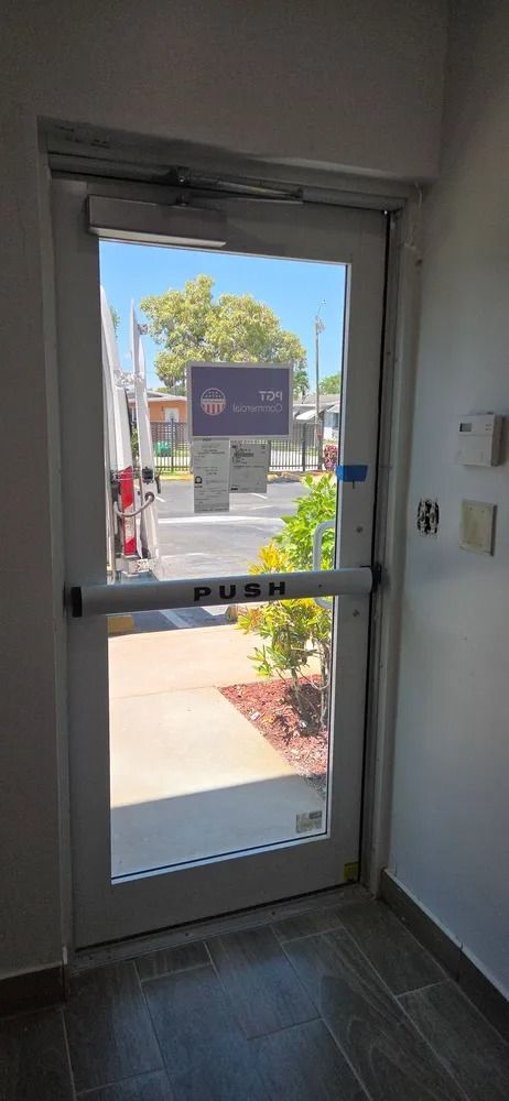 View through a glass exit door to a sunny outdoor walkway and sign, with a small plant bed outside