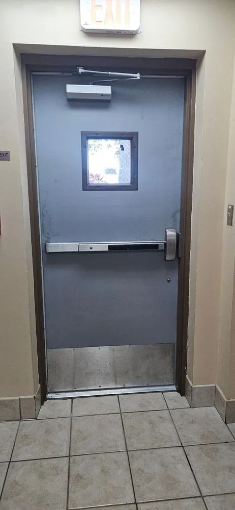 Gray metal door with small window and push bar in a tiled hallway, with an exit sign above.