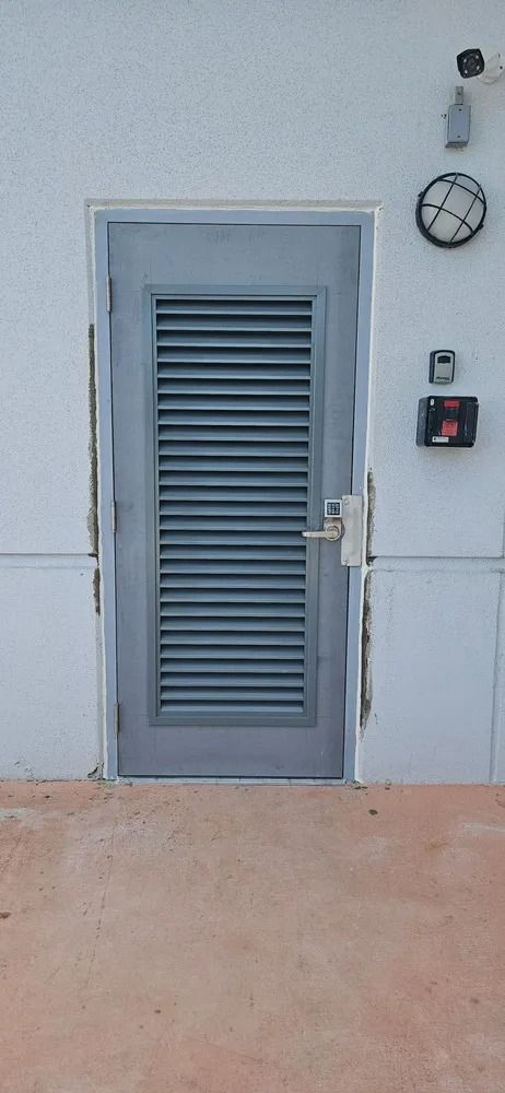 Blue-gray door with louvered panel, wall clock, and red alarm box on a white exterior wall