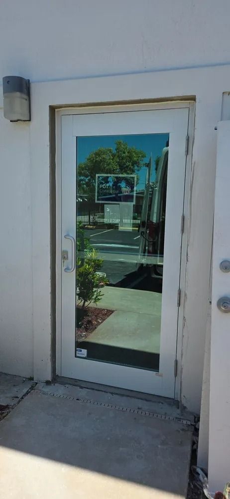 White-framed glass door in a stucco wall, reflecting a sunny street and trees.