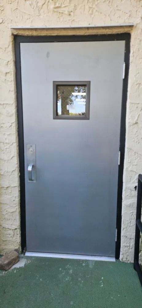 Gray exterior door with small window and handle, set in a beige wall above green flooring