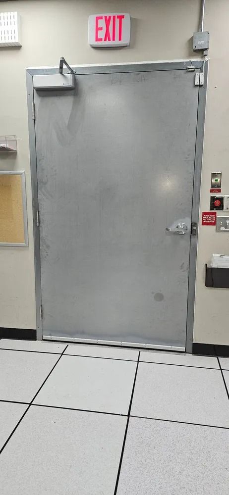 Gray metal emergency exit door with push bar and illuminated EXIT sign above it in a tiled hallway.