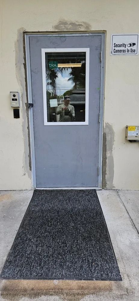 Gray door with a window at a building entrance, with a dark doormat on the concrete sidewalk.