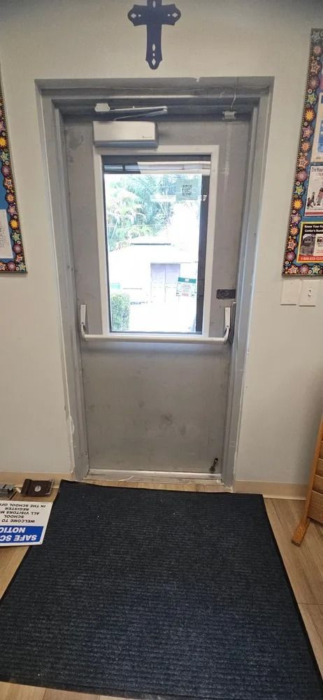 Indoor door with a small window, cross above it, and daylight visible outside; dark floor mat in front