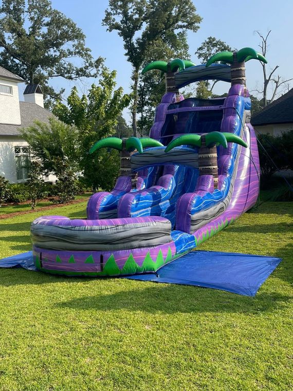 Purple-and-green inflatable water slide on a lawn beside a house, with blue mats at the base.