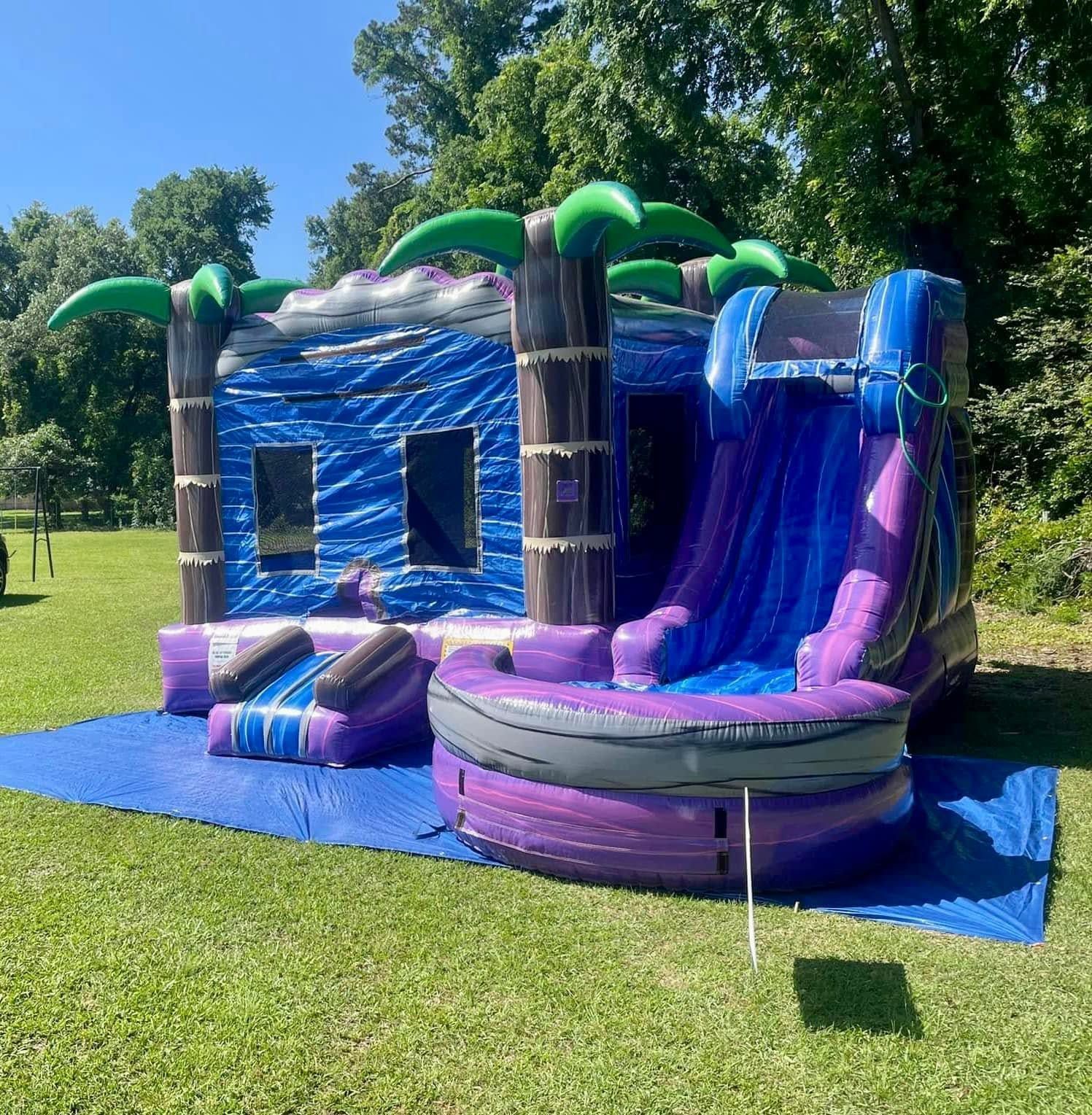 Purple-and-blue tropical inflatable bounce house with slide on a grassy lawn in a sunny park