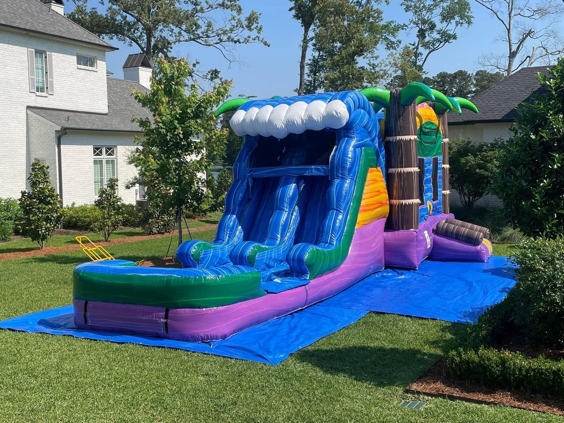 Blue and purple inflatable water slide in a suburban yard, set up on a lawn near houses.
