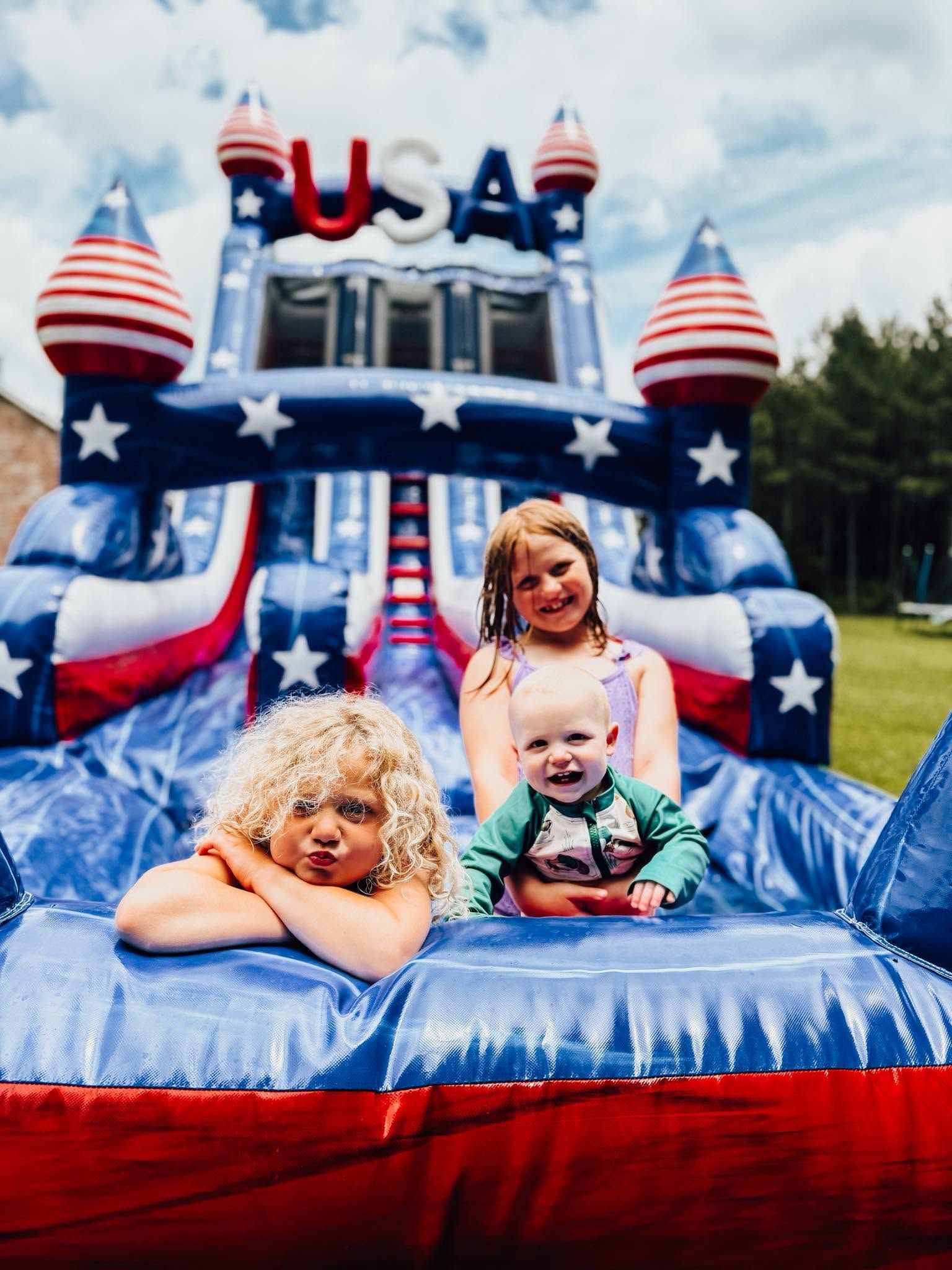 Three children on a blue-and-red patriotic inflatable bounce house with USA stars and stripes decorations
