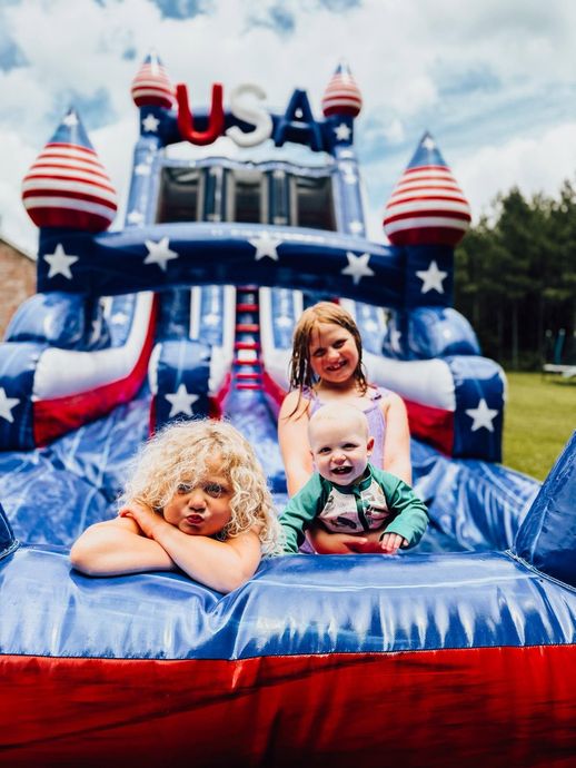 Three children on a blue-and-red patriotic inflatable bounce house with USA stars and stripes decorations