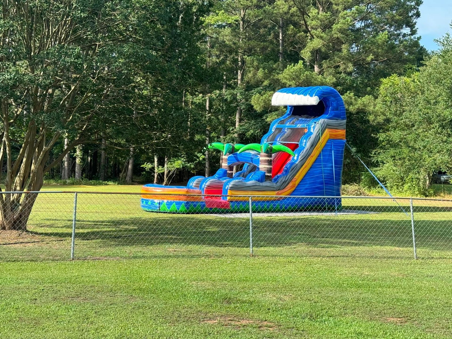 Colorful inflatable water slide and bounce house in a grassy park, with trees and a fence in front